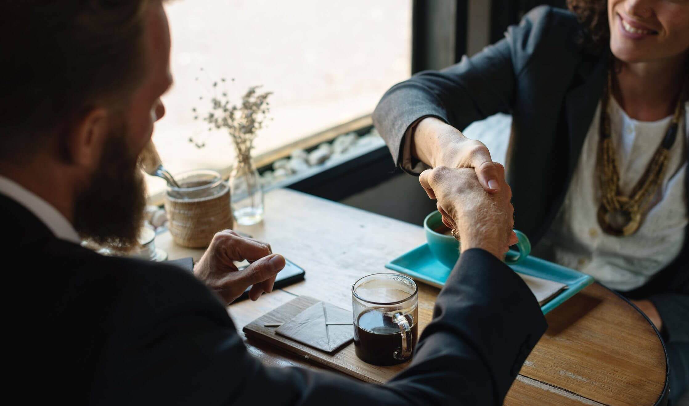 Two business people shake hands while sitting down at a coffee shop table. 