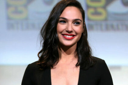 Actress Gal Gadot wears a dark top and smiles wearing red lipstick.