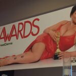 A brunette woman wearing a revealing orange dress lies on her side with an gold awards trophy next to the text AVN Awards 2019.