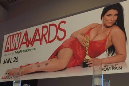 A brunette woman wearing a revealing orange dress lies on her side with an gold awards trophy next to the text AVN Awards 2019.