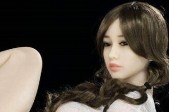 A pale skinned sex doll poses on its side in a white see-through top and short black skirt.