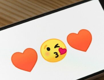 A smartphone shows two heart emojis with a yellow-face kiss emoji in the middle.