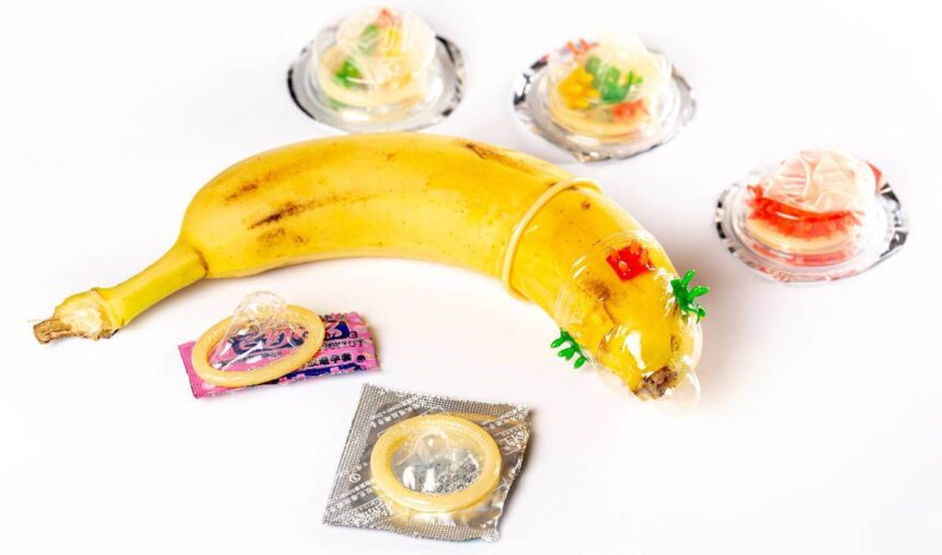 A banana is shown weraing a condom with various unopened condoms lying around it.