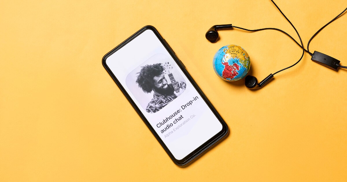 Clubhouse Invite-only Audio Chat App Open in Mobile Phone with a Globe and Earphone Display in Background