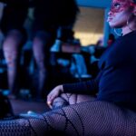 A dancer in fishnet stockings sits in front of a lighted mirror while other coworker getting ready