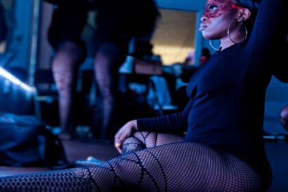 A dancer in fishnet stockings sits in front of a lighted mirror while other coworker getting ready