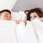 A couple peer at each other from under the bed covers.