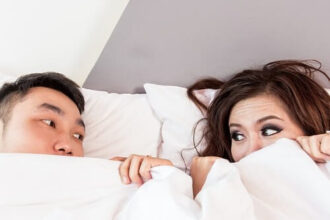 A couple peer at each other from under the bed covers.