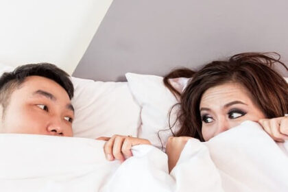 A couple peer at each other from under the bed covers.