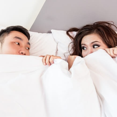 A couple peer at each other from under the bed covers.