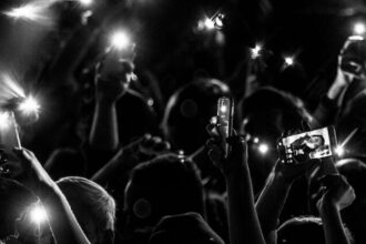 A black and white photo of a crowd at a concert or event. Everyone has their hands raised and most are holding cell phones. The phones have their flashlight setting turned on.
