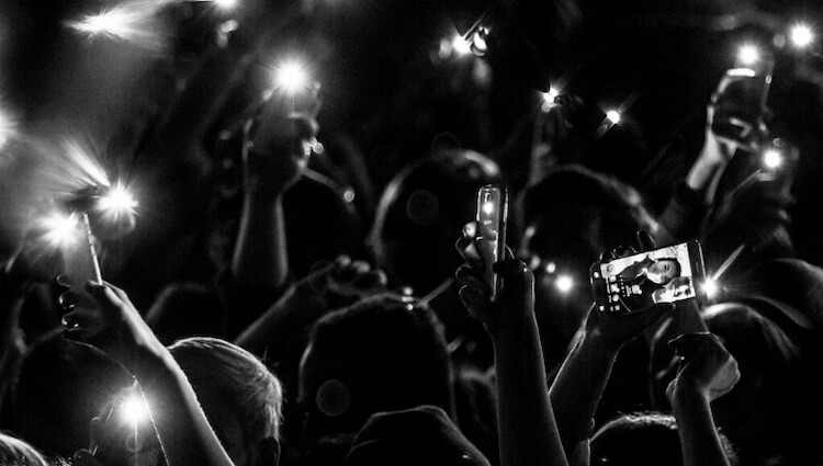 A black and white photo of a crowd at a concert or event. Everyone has their hands raised and most are holding cell phones. The phones have their flashlight setting turned on.