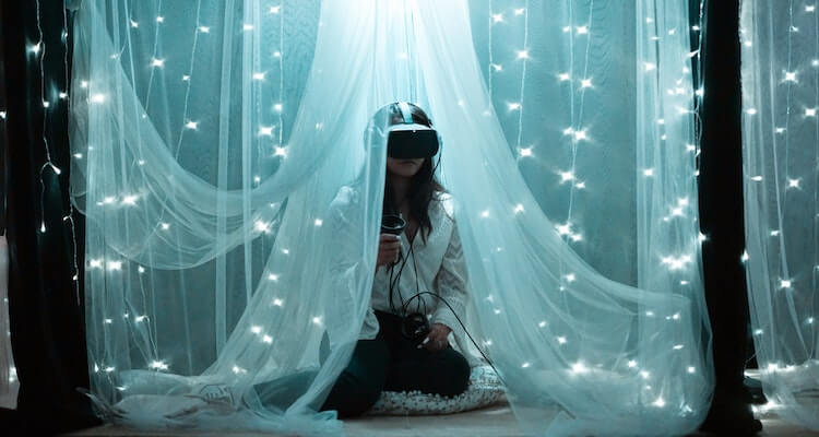 A decorative image of a woman sitting in a canopy wearing a virtual reality headset. She's surrounded by pillows and twinkly lights.