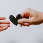Decorative image of two hands reaching towards each other. On one, you can see a wedding ring. The other is holding a black paper heart.
