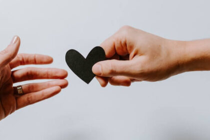 Decorative image of two hands reaching towards each other. On one, you can see a wedding ring. The other is holding a black paper heart.