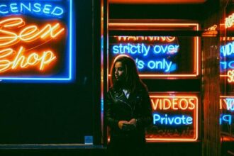 A woman in a leather jacket standing outside a storefront at night. It's dark all around, but you can see the neon signs with sayings like "Licensed Sex Shop", "Strictly over 18s only" and "private videos".