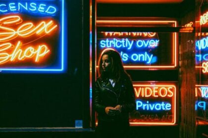 A woman in a leather jacket standing outside a storefront at night. It's dark all around, but you can see the neon signs with sayings like "Licensed Sex Shop", "Strictly over 18s only" and "private videos".