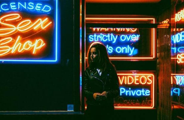 A woman in a leather jacket standing outside a storefront at night. It's dark all around, but you can see the neon signs with sayings like "Licensed Sex Shop", "Strictly over 18s only" and "private videos".