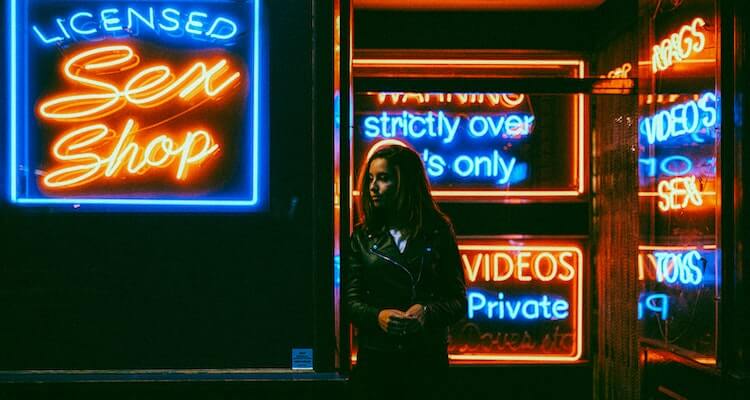 A woman in a leather jacket standing outside a storefront at night. It's dark all around, but you can see the neon signs with sayings like "Licensed Sex Shop", "Strictly over 18s only" and "private videos".