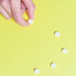 Light yellow background with a trail of small round white pills moving up the center of the image. There's a light-skin human hand reaching into frame from the top-left corner grabbing one of the pills.