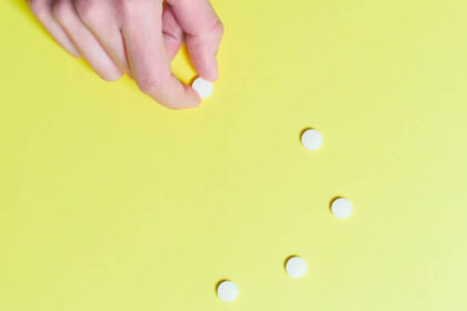 Light yellow background with a trail of small round white pills moving up the center of the image. There's a light-skin human hand reaching into frame from the top-left corner grabbing one of the pills.