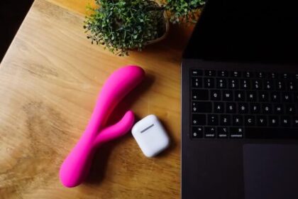 A pink rabbit vibrator and AirPods case laying on a table next to an open laptop.