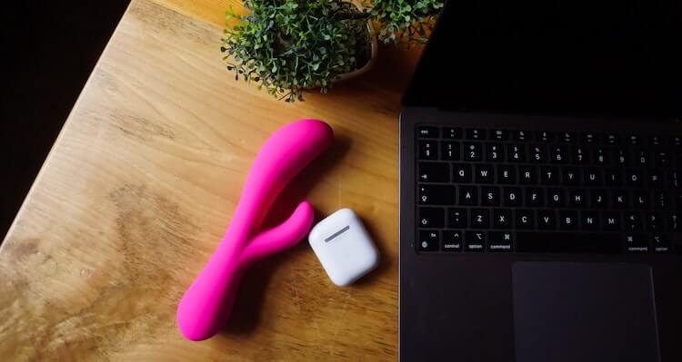 A pink rabbit vibrator and AirPods case laying on a table next to an open laptop.