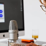 The view of standing behind someone at their desk. This person is wearing headphones and speaking into a recording microphone. You can also see the keyboard and monitor, with the screen showing blurred out audio editing software.