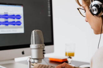 The view of standing behind someone at their desk. This person is wearing headphones and speaking into a recording microphone. You can also see the keyboard and monitor, with the screen showing blurred out audio editing software.