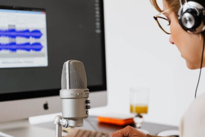 The view of standing behind someone at their desk. This person is wearing headphones and speaking into a recording microphone. You can also see the keyboard and monitor, with the screen showing blurred out audio editing software.