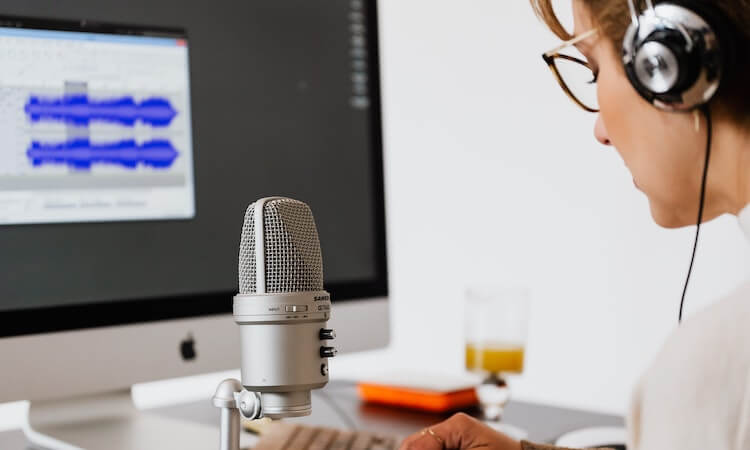 The view of standing behind someone at their desk. This person is wearing headphones and speaking into a recording microphone. You can also see the keyboard and monitor, with the screen showing blurred out audio editing software.