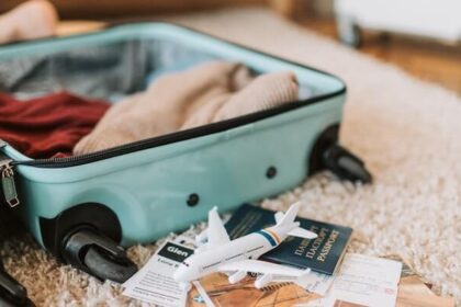 A suitcase open on the floor mid-packing. You can see sweaters inside the suitcase and a passport and model airplane next to it.