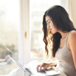 Woman looking at a laptop in a bed. You can see a blurry European city outside of the window.