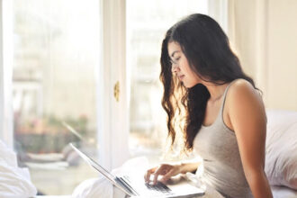 Woman looking at a laptop in a bed. You can see a blurry European city outside of the window.