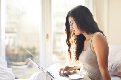 Woman looking at a laptop in a bed. You can see a blurry European city outside of the window.