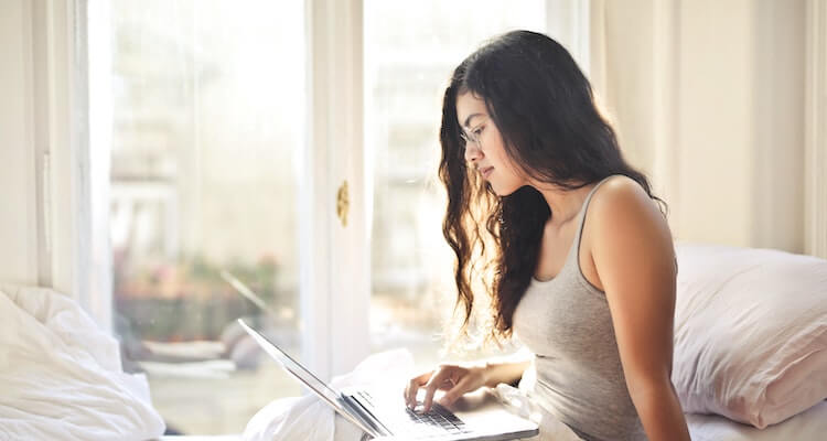 Woman looking at a laptop in a bed. You can see a blurry European city outside of the window.